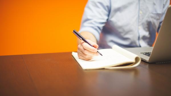 Man writes in notebook next to his laptop in front of an orange wall