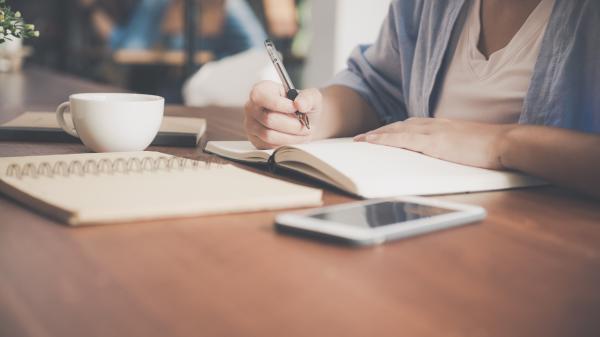 Woman writes in notebook with other books and a teacup on the table in front of her
