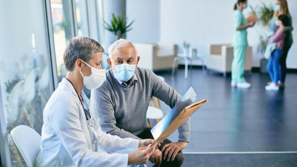 Female health practitioner sits talking to an older man with a mask 
