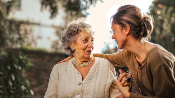Sitting outside, a younger woman clasps the hand of an older woman as they smile at one another