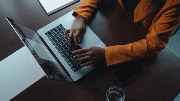 We look down on a woman in an orange shirt typing on her laptop