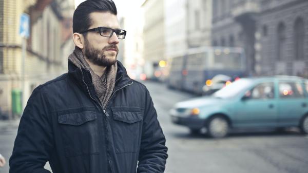 On a street a contemplative man with thick glasses stands with his hands in his coat pockets