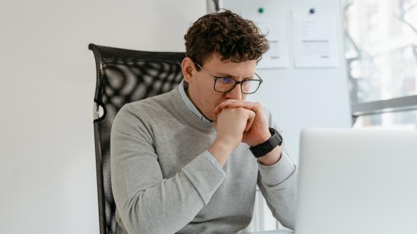 A curly-haired young man clasps his hands while looking at his laptop screen