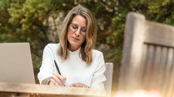 At an outside table a woman with brown hair focuses on writing something