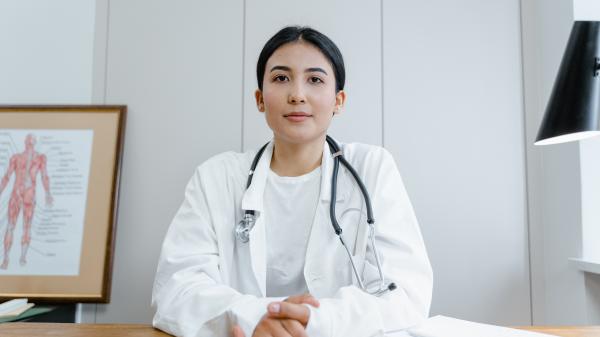 A woman wearing a stethoscope and white coat sits behind a desk, observing the camera