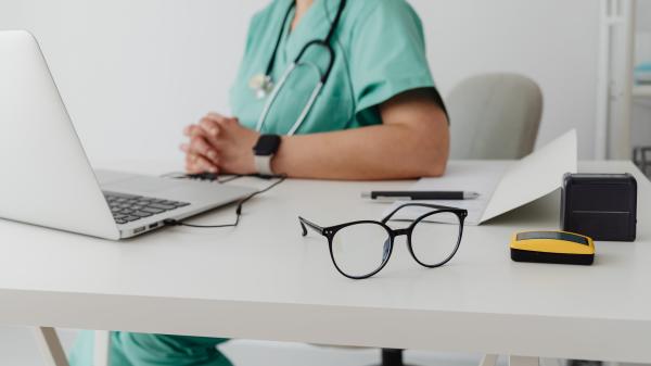 A person in scrubs sits behind their desk with a laptop and glasses in front of them