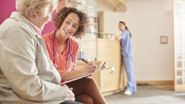 A woman with a clipboard sits with an older woman in what appears to be a medical setting