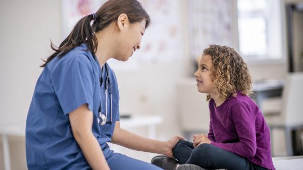 Woman in blue scrubs looks calmly at boy in purple sitting on bed