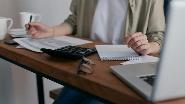 A desk is filled with a laptop, calculator and papers as a woman takes notes