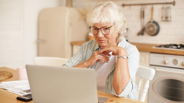An older woman has her hand beneath her chin while she looks at a laptop screen