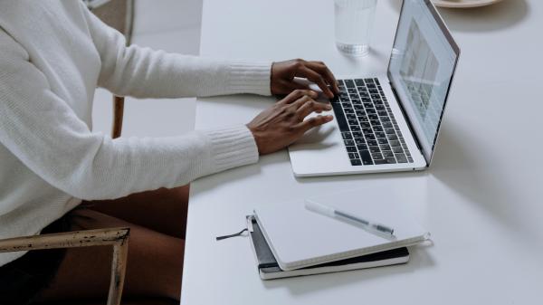 On a white desk, a woman's arms lean as she types on her laptop, a notebook beside her
