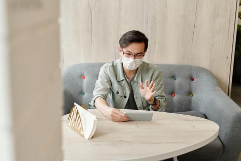 Young man sits on a couch with a mask on waving to a person on his phone in front of him