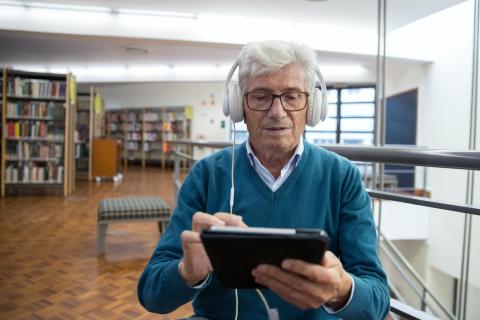 Older man sits in a library using a tablet with white headphones on
