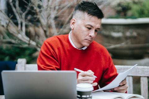 Man in red jumper reads with a laptop in front of him
