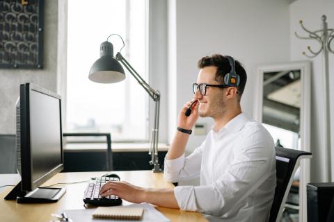 Man with a headset sits at a home office speaking on the phone while on the computer