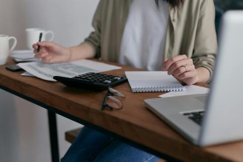 A desk is filled with a laptop, calculator and papers as a woman takes notes