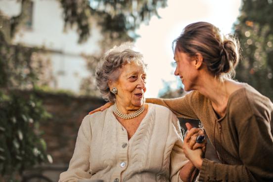 Sitting outside, a younger woman clasps the hand of an older woman as they smile at one another